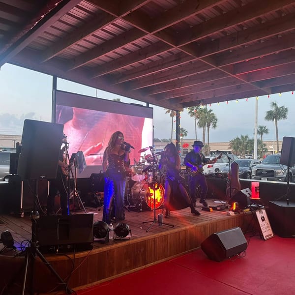 A band performs on an outdoor stage at Texas Huddle, with palm trees and cars in the background.