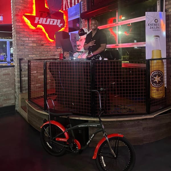 A DJ smiles behind a booth with a red neon Texas Huddle sign and an Estrella Jalisco bike.