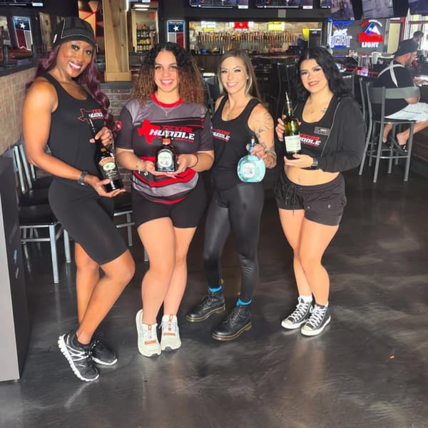 Four women in Texas Huddle branded attire holding liquor bottles inside the restaurant.
