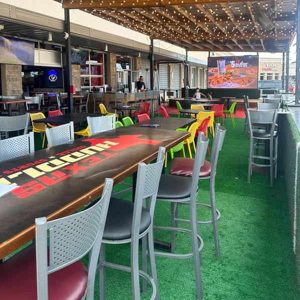Outdoor patio at Texas Huddle with long tables, colorful chairs, string lights, and a large TV screen.