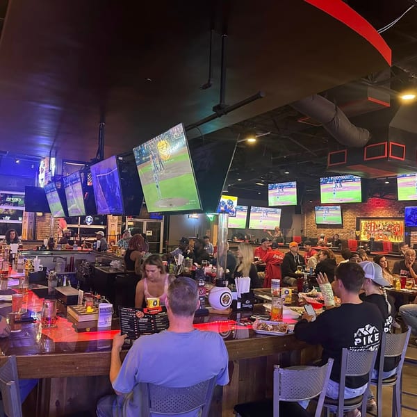 Patrons at Texas Huddle bar watching sports on multiple large screens, with drinks and food.